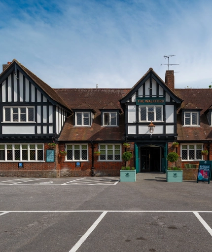 The exterior facade, signage, seating area, and car park of The Walkford in Walkford, with flower baskets hanging on the walls, string lights above the seating area, and accessible parking spaces near the entrance.