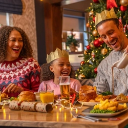 A festive Christmas meal taking place beside a decorated Christmas tree adorned with red and gold baubles, candy cane ornaments and warm fairy lights. Three people seated at a dining table wear paper crowns from Christmas crackers while enjoying a traditional festive meal. One person pours gravy from a gravy boat onto a dinner plate. The table is set with roast dinners, vegetables, glasses of orange juice, a pint of beer, Christmas crackers and string lights, creating a warm and seasonal restaurant setting.