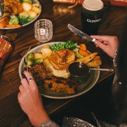 A close up view of a nut roast dinner served on a plate which sits on a table next to a glass of Guinness, a glass of champagne, a Christmas cracker, and another plate of food. A person sitting at the table is holding a knife and fork above the plate.