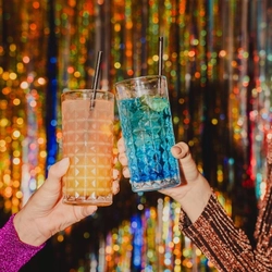 Two hands raise colourful cocktails in a celebratory toast in front of a glittering gold and silver tinsel backdrop. One drink is a bright blue cocktail with lime and ice, while the other is a pink-orange cocktail served over ice, both in textured glass tumblers with black straws. The festive close-up highlights sparkling New Year’s Eve party decorations and vibrant drinks.