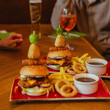 Two stacked burgers topped with festive garnishes are served with chips, onion rings and gravy on red plates at a wooden table. Glasses of white wine, rosé and beer are arranged around the table, with diners seated nearby. The cosy pub setting features warm lighting and festive decorations in the background.