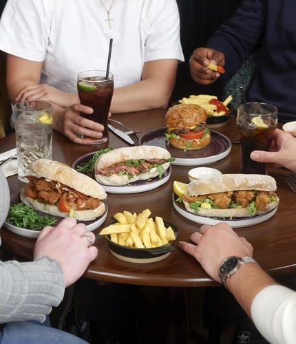 A group of four people seated around a small round wooden table, sharing a meal. The table is filled with plates of food, including baguette-style sandwiches, a burger, and several small bowls of chips. There are also drinks on the table, including glasses of soda with straws and a cup of coffee. Only the individuals’ torsos and arms are visible as they sit closely together, reaching toward the food.