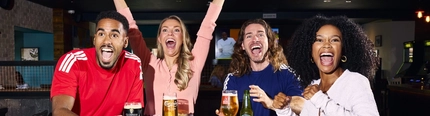 An image showing 4 friends at a table enjoying drinks and celebrating while watching on a football match within the interior seating area at a Flaming Grill venue.