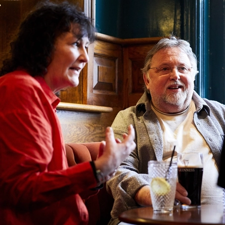 Two people sitting at a table inside a pub. One person is holding a glass of Guinness which sits on the table next to a glass of soft drink.