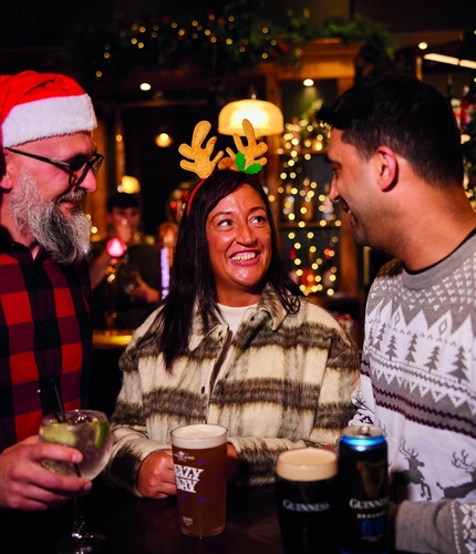 Three people are standing at a tall table with drinks,  in a festive, warmly lit bar. Two of them are wearing Christmas themed headwear.  The background features Christmas decorations, including garlands and lights, as well as hanging glasses above the bar area.