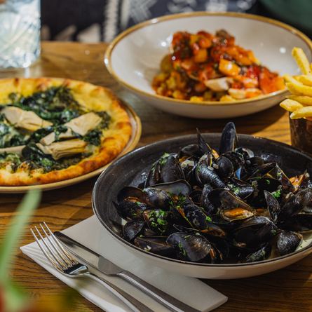 An advertising image showing a selection of main dishes from the Prix Fixe menu sat on a table within the interior restaurant seating area at The Four Oaks.