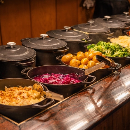 Cauliflower cheese, red cabbage, new potatoes, broccoli, mashed potato, carrots, and roast potatoes, each served in large roasting dishes which sit on a carvery counter.