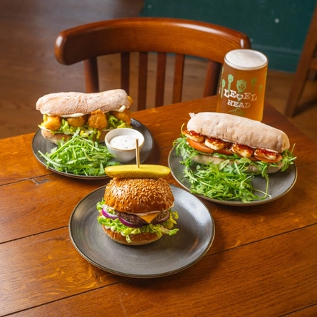 A Cheeseburger, a Cod Goujon Ciabatta, and a Halloumi Ciabatta, each served on a plate which sits on a wooden restaurant table along with a glass of Level Head.