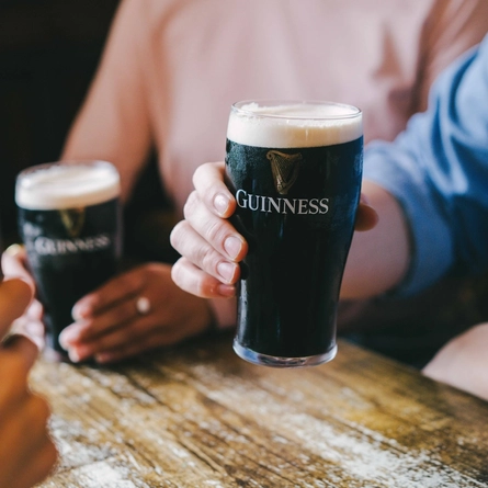Three people are holding pints of Guinness beer, seated at a wooden table. The focus is on the hands and the glasses.