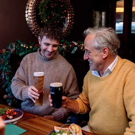 An image focusing on 2 people sat with others at a table within the interior restaurant seating area celebrating with a cheers while food dishes from the Festive Fayre menu at a Community Social venue.