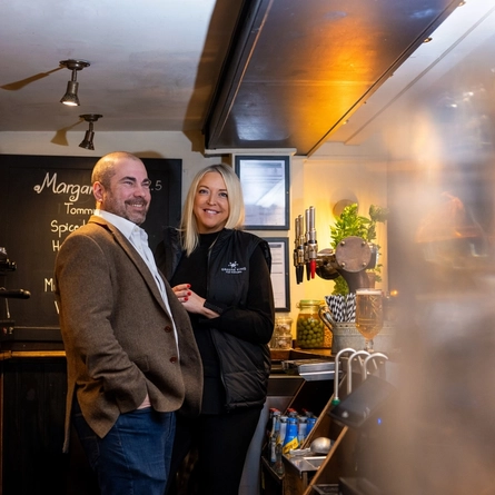 A Man and Women stood behind a bar smiling 