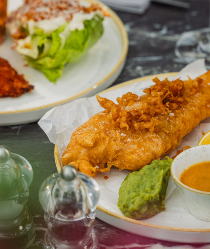 A plated fish, chips and peas dish with a Chicken Kyiv dish in the background sat on a table within the exterior beer garden seating area at a Crafted venue.