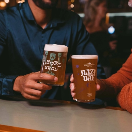 A close up view of two people sitting at a table in a bar; one of them holding a glass of Level Head; the other holding a glass of Hazy Day. A Christmas tree is visible in the background.