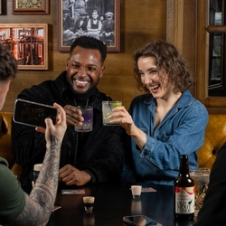Four people sit around a dark wooden table in a pub setting. Two people on the far side of the table hold drinks and are smiling while looking toward a phone being held up by a person in the foreground. The person next to the phone holder also looks toward the group. Various drinks are on the table.