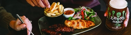 A close up view of sliced chicken served on a plate with salad and a bowl of fries. The plate sits on a wooden table along with a glass of Level Head and another plate of food; and two people are sitting at the table, one of them picking up one of the fries and holding the glass of Level Head.