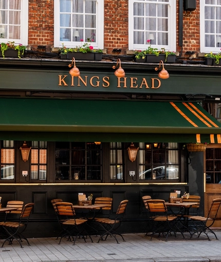 The exterior facade, signage, and seating area at the King's Head in Bayswater, with a dark green awning hanging over the wooden tables and chairs; and flower boxes beneath the upper windows.