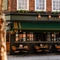 The exterior facade, signage, and seating area at the King's Head in Bayswater, with a dark green awning hanging over the wooden tables and chairs; and flower boxes beneath the upper windows.