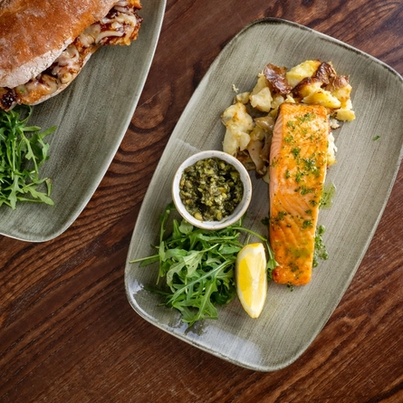 A bird's eye view of a sub sandwich served on a plate with salad leaves; and a piece of salmon served on a plate with salad leaves, dipping sauce, and a slice of lemon. The two plates sit on a wooden restaurant table.