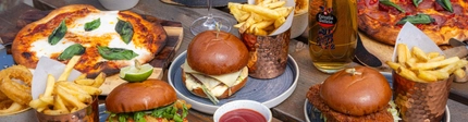 An advertising image of a selection of burger and pizza dishes with fries and onion rings and a selection of drinks sat on a table within the exterior beer garden seating area at a Crafted venue.