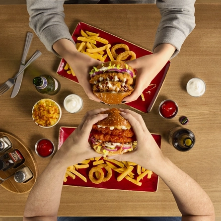 The hands of two people seated at a wooden table each holding a burger.  On teh table in front of each person are plates of fries and onion rings.