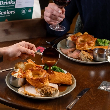 A table setting inside a cozy restaurant or pub, where two people are seated and sharing a meal. The table holds plates of food, including what appears to be a roast dinner, along with wine glasses, cutlery, and condiments. One person reaches toward the food while the other sits opposite. .