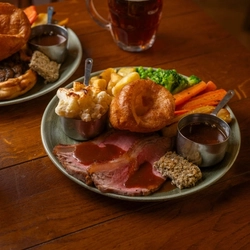 A Roast Beef dinner served on a plate and featuring Yorkshire pudding, stuffing, cauliflower cheese, roast potatoes, vegetables, and gravy. The plate sits on a wooden restaurant table along with two glasses of beer and another plate of food.