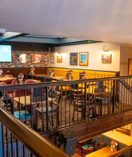 A view from the balcony of another upper floor restaurant seating area inside the Old School House, with a long, upholstered booth seat along one side of the room, framed artwork on the wall above it, and a TV on another wall.