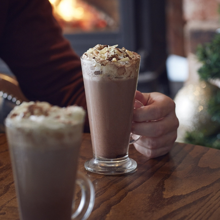 An advertising image of a hot chocolate topped with cream being enjoyed within the interior restaurant seating area at a Chef & Brewer venue.