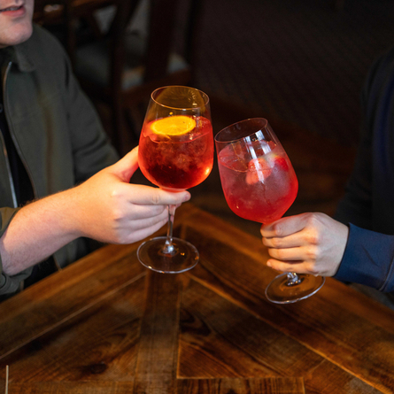 A close up view of two people at a wooden table, holding cocktails in wine glasses and clinking them together.