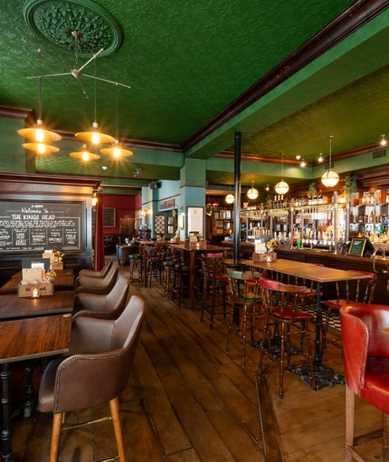 The bar and interior restaurant seating area at the King's Head, with dark wood floors; green, textured wallpaper on the ceiling; and a long, red, upholstered booth seat along one wall by the window.