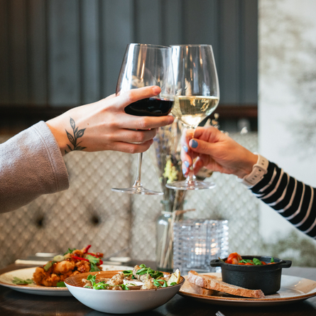 Two people at a restaurant table clinking together glasses of red and white wine. On the table are three meals served on two plates and a bowl. 