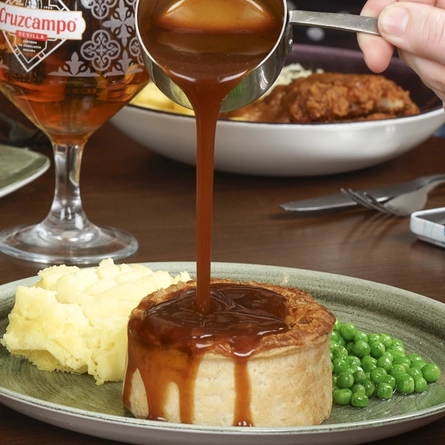 A round table filled with main dishes, including a burger with fries, a pie with mash and peas. Several drinks—such as beer, rosé wine, and a bottle of cider —are placed around the table. The hands and arms of multiple people seated together are visible as they reach toward their food and raise their drinks.
