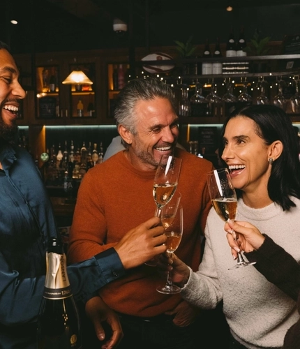 Four people standing at a table in a bar, laughing together and raising glasses of champagne. A bottle of Lanson champagne stands on the table, and a Christmas tree decorated in red and gold stands beside them.