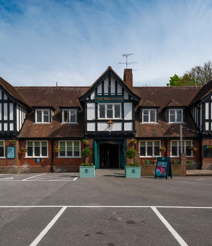 The exterior facade, signage, seating area, and car park of The Walkford in Walkford, with flower baskets hanging on the walls, string lights above the seating area, and accessible parking spaces near the entrance.