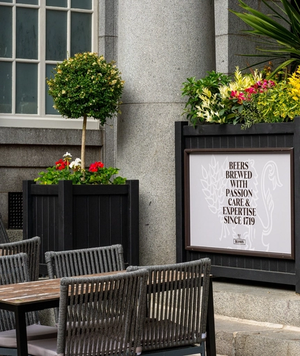 The exterior beer garden seating area at the Old School House, with large stone pillars, and a topiary tree in a wooden planter box next to the tables and chairs.