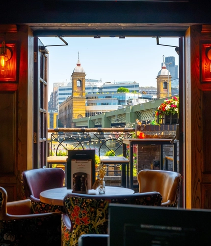 An interior restaurant seating area at The Anchor Bankside in Southwark; with upholstered chairs, wall lights on the wood panelled walls, and open doors leading out into the beer garden overlooking the River Thames.