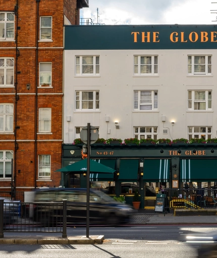 A view from across the busy street of the exterior facade, signage, and seating area of The Globe in Marylebone; with large green awnings hanging above the seating area.