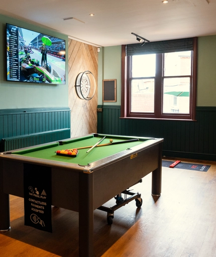 The interior games area at The Richmond Arms in Bournemouth, with a pool table; a dartboard; a TV on the wall; and bar stool seating.