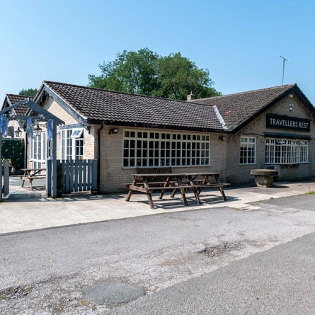 An image showing the exterior facade of the venue and outdoor seating with wooden benches at The Travellers Rest.