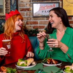 Three people seated around a festive pub table enjoying food and drinks. One person wearing a red paper crown and red jumper holds a pint of beer, while another in a green blouse holds a glass of cola garnished with a dried citrus slice. Plates of seafood dishes with salad and lemon wedges are arranged on the table alongside green Christmas crackers, with garlands, fairy lights and a brick wall creating a warm seasonal pub setting.