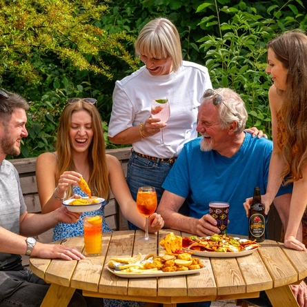 A mixed group of people seated around a wooden table, in a beer garden, enjoying a variety of sharers and drinks.