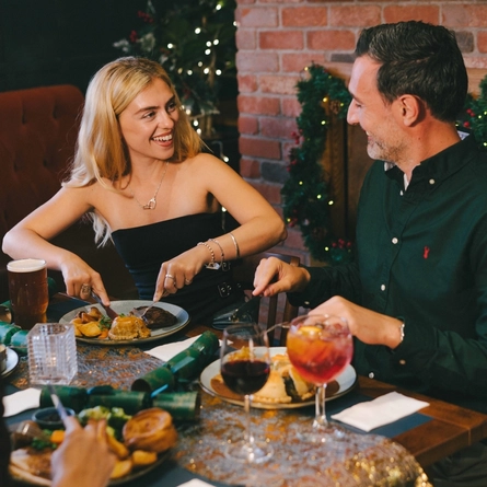 An image focusing on 2 people sat with friends enjoying various main dishes and a selection of drinks while sat within the interior restaurant seating area.