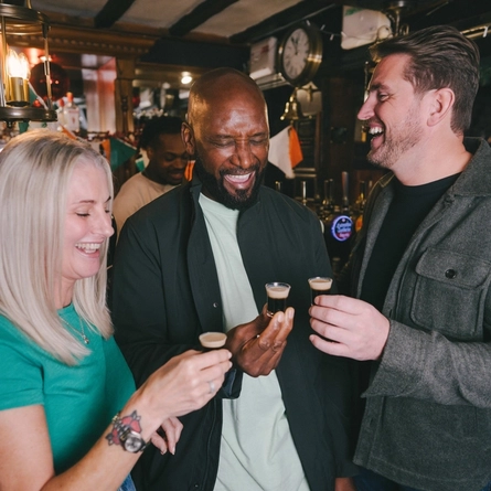 Three people standing at the bar inside a pub, holding shots of Guinness.