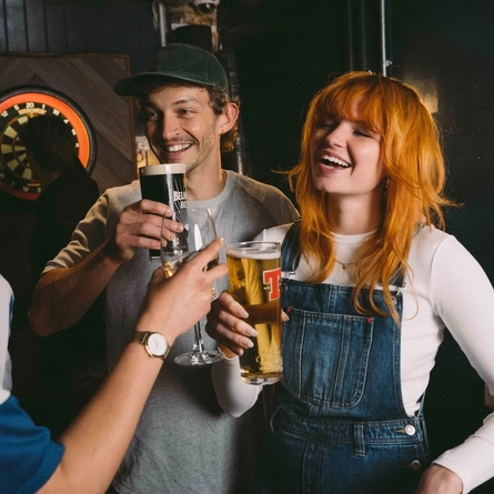 Three students standing in front of a dartboard inside a pub; one is holding a glass of wine, another is holding a glass of stout, and the third is holding a glass of Tennent's.