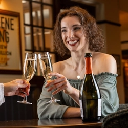 Two women seated at an indoor table.  They are both holding champagne flutes containing prosecco in a toast.  A bottle is on the table between them.