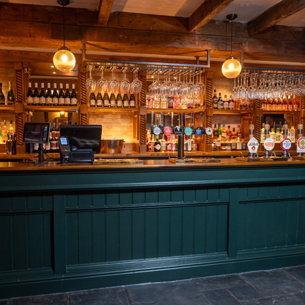 A close up view of the bar inside The Rising Sun in Wistaston, with stone flooring, wooden beams on the ceiling, and wine glasses hanging in racks above the counter.