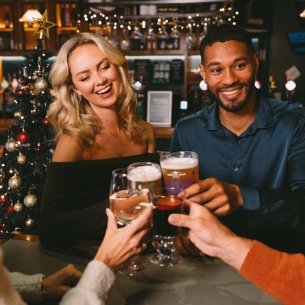 Four people at a table in a bar, clinking together glasses of beer, white wine, and mulled wine. A Christmas tree decorated in red and gold, and lit with fairy lights stands behind the table.