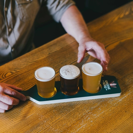 A man is seated at an indoor wooden table which is holding a Beer Flight Paddle with 3 mini pint glasses of beer on it. The man's arms are on the table and he is reaching for one of the glasses.