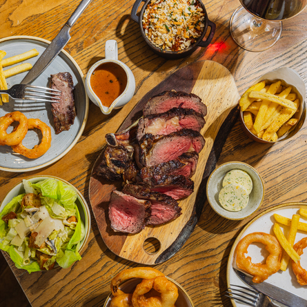 An advertising image showing a boarded Ribeye Sharer dish including onions rings, chips, salad and sauce sat on a table within the interior restaurant seating area at The Four Oaks.