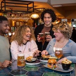 Four people stand and sit around a bar-height table in a busy pub. The table is covered with food, including sliders, fries, dips, and small plates. Several drinks are visible, such as a pint of beer, a canned drink, and another beer in a stemmed glass. One person standing behind the table holds two pints.